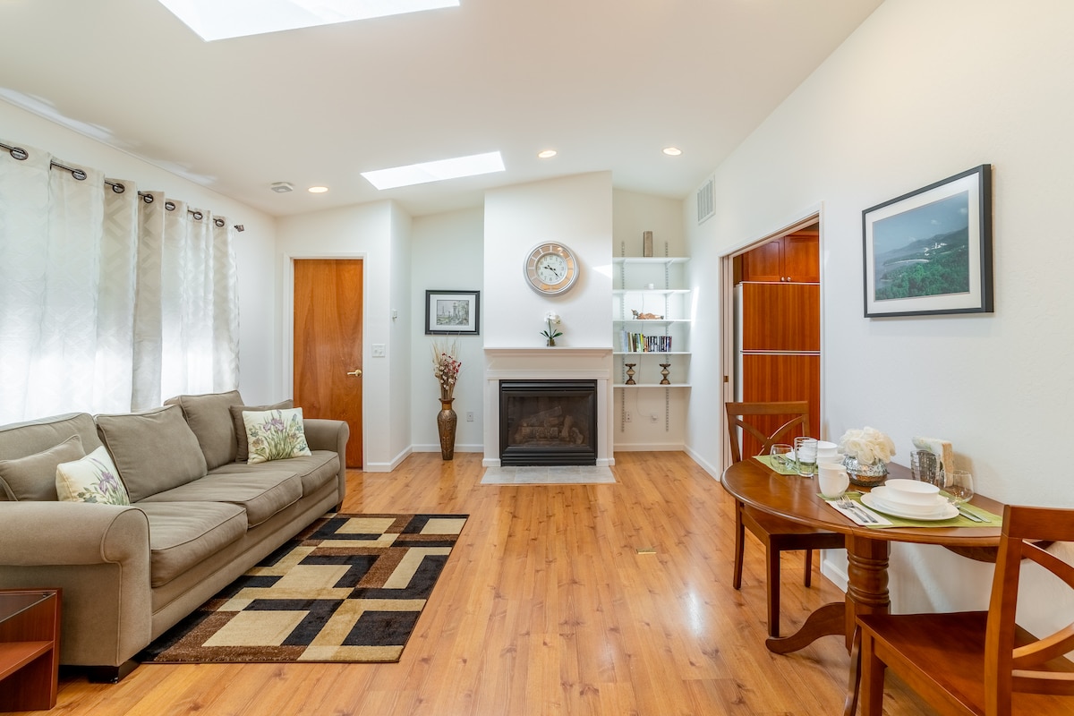 A well-lit living area features a comfortable sofa and a decorative area rug. Natural light enters through skylights and windows adorned with sheer curtains. A fireplace is centered against the wall, complemented by a dining table set for two, creating an inviting atmosphere.