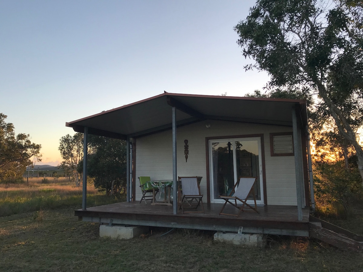 A wooden deck is featured in front of the bungalow, with two white chairs and a small table positioned for outdoor relaxation. The structure is surrounded by green trees and open fields, under a clear sky transitioning to sunset hues.