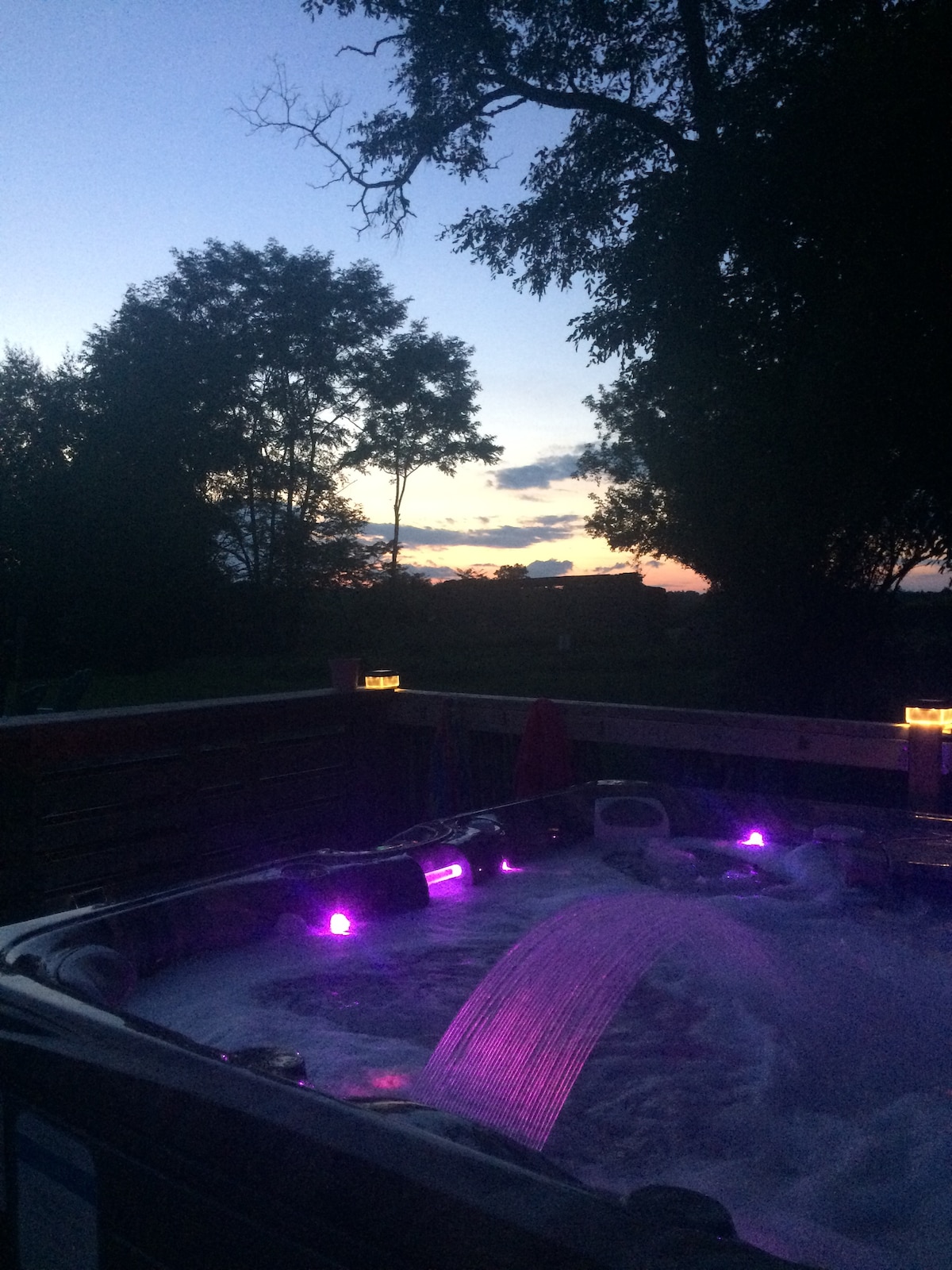 A hot tub is positioned on a wooden deck, surrounded by lush trees and facing a tranquil sunset sky with hues of blue and orange. Soft purple lights illuminate the water, creating a serene ambiance complemented by subtle hints of natural scenery.