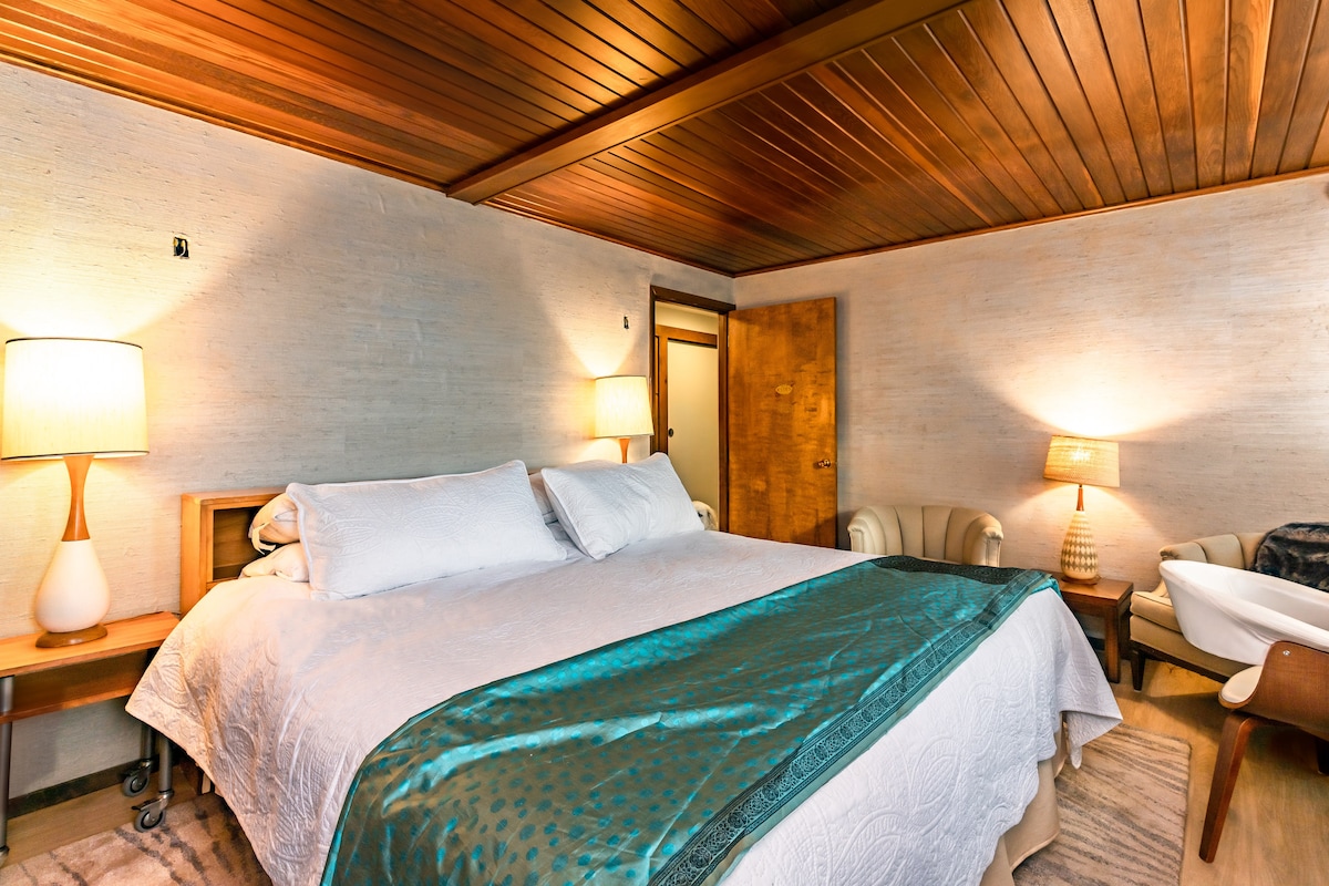 A spacious bedroom is furnished with a king bed, complemented by a decorative bedspread in teal. Two bedside lamps provide soft lighting beside the bed. A redwood ceiling and grasscloth wallpaper enhance the room's natural aesthetic.