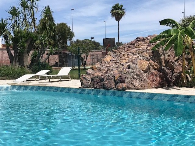 A clear blue swimming pool reflects sunlight, bordered by a stone formation and lush greenery. Two lounge chairs are positioned nearby, providing a space for relaxation. In the background, sports courts are visible, enhancing the recreational atmosphere.