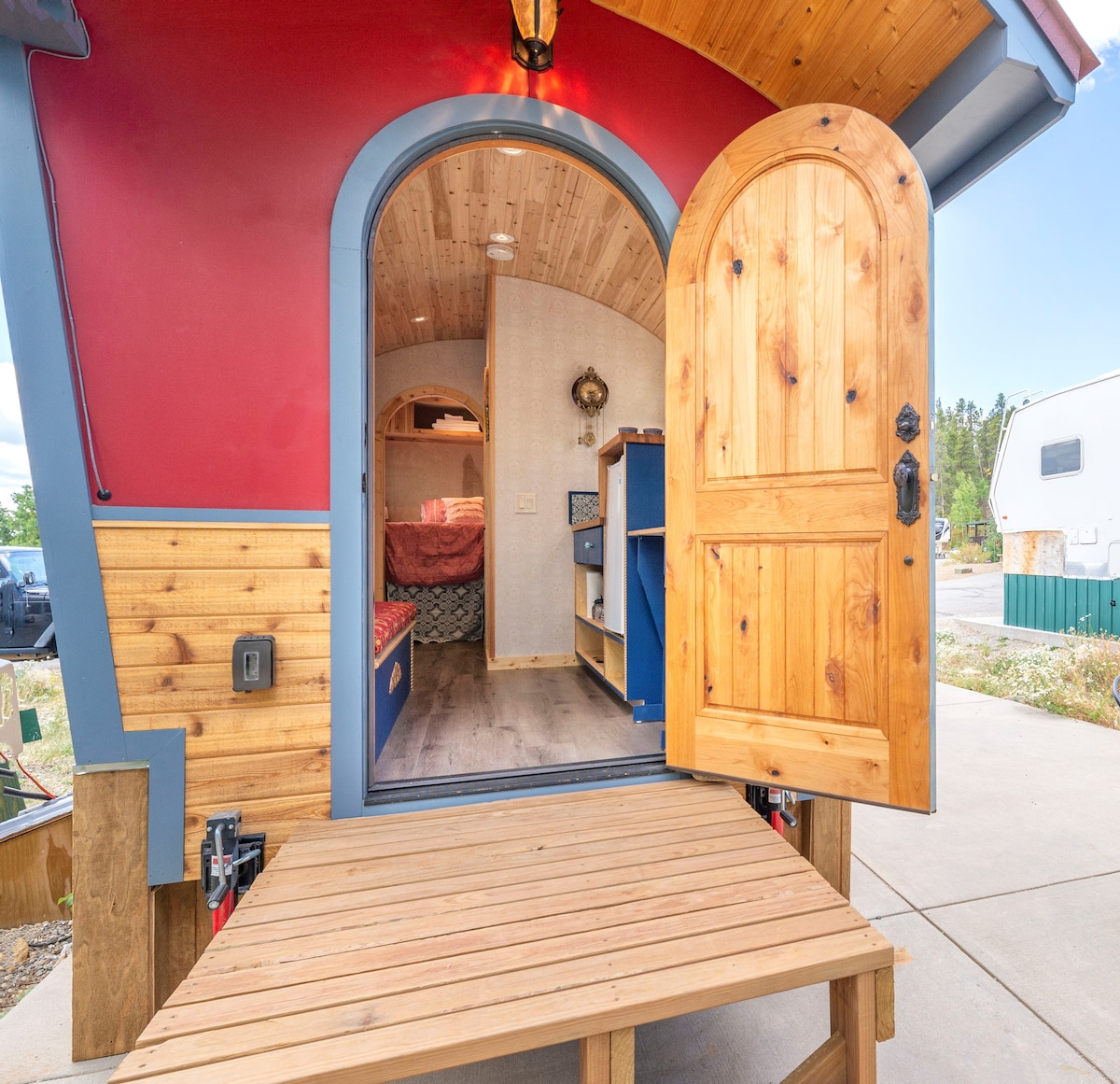 A welcoming entryway is shown, featuring a wooden door opened to reveal the interior of a colorful gypsy wagon. Natural wood accents can be seen inside, while a cozy sleeping area is visible in the background, complemented by decorative shelving and warm lighting.
