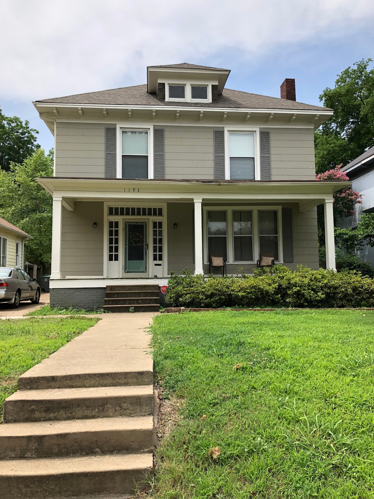 A charming turn-of-the-century home is presented, featuring a light gray exterior with white trim. A set of stairs leads to a spacious front porch, furnished with two rocking chairs, and a well-maintained garden surrounds the entrance. The roof is adorned with gables, adding character to the structure.