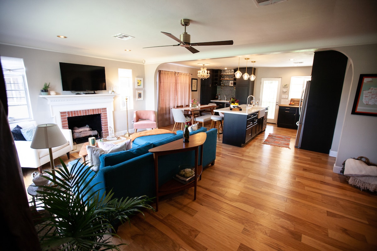 A spacious living area features hardwood floors and a cozy seating arrangement with a plush blue couch. A flat-screen TV sits above a classic fireplace, and an open kitchen with a large island and modern appliances is visible in the background, providing a bright and inviting atmosphere.