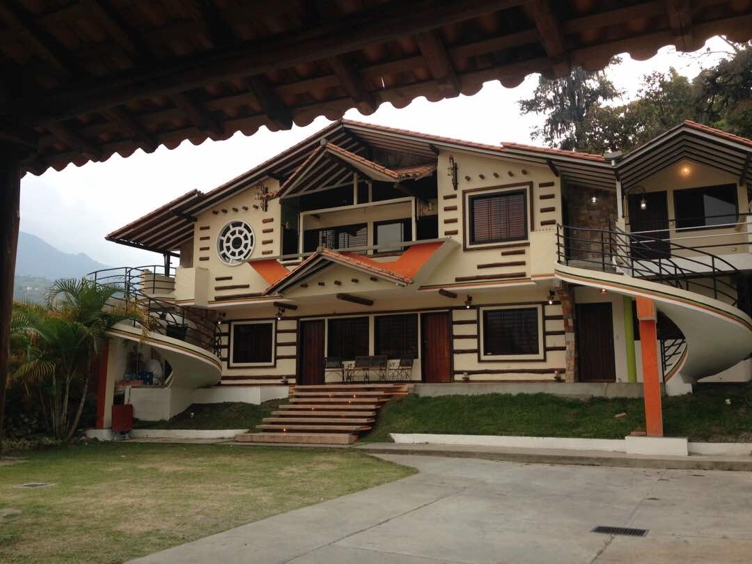 A spacious cabin structure with an artistic exterior, featuring multiple balconies and large windows. The building is surrounded by grassy areas and palm trees, with a covered terrace offering seating. The backdrop consists of rolling hills, enhancing the attractive setting.