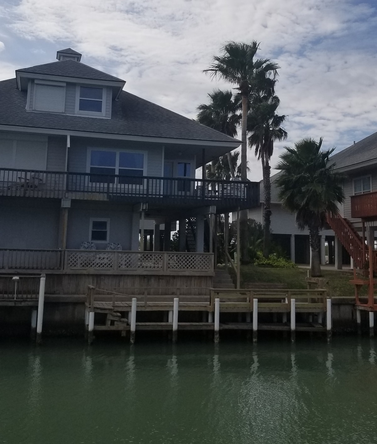 The exterior of a sea cottage is displayed, featuring a multi-level design with a large balcony and palm trees nearby. A wooden dock extends into the water, complemented by a grassy area. The sky is partly cloudy, reflecting a serene atmosphere by the water.