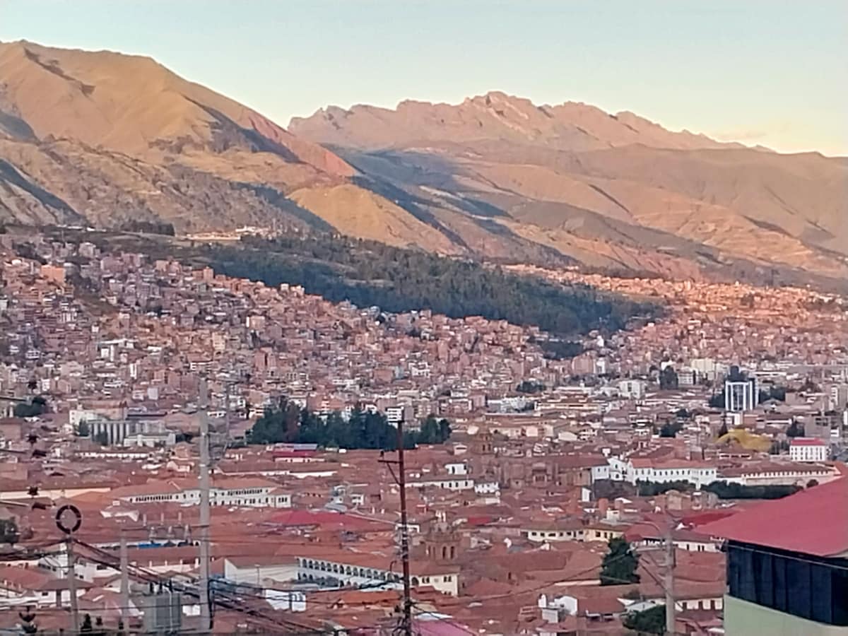 A panoramic view of Cusco displays a sprawling city with terracotta rooftops, nestled in a valley surrounded by majestic mountains. The landscape features varying elevations and a gradient of colors from the setting sun illuminating the peaks in the distance.