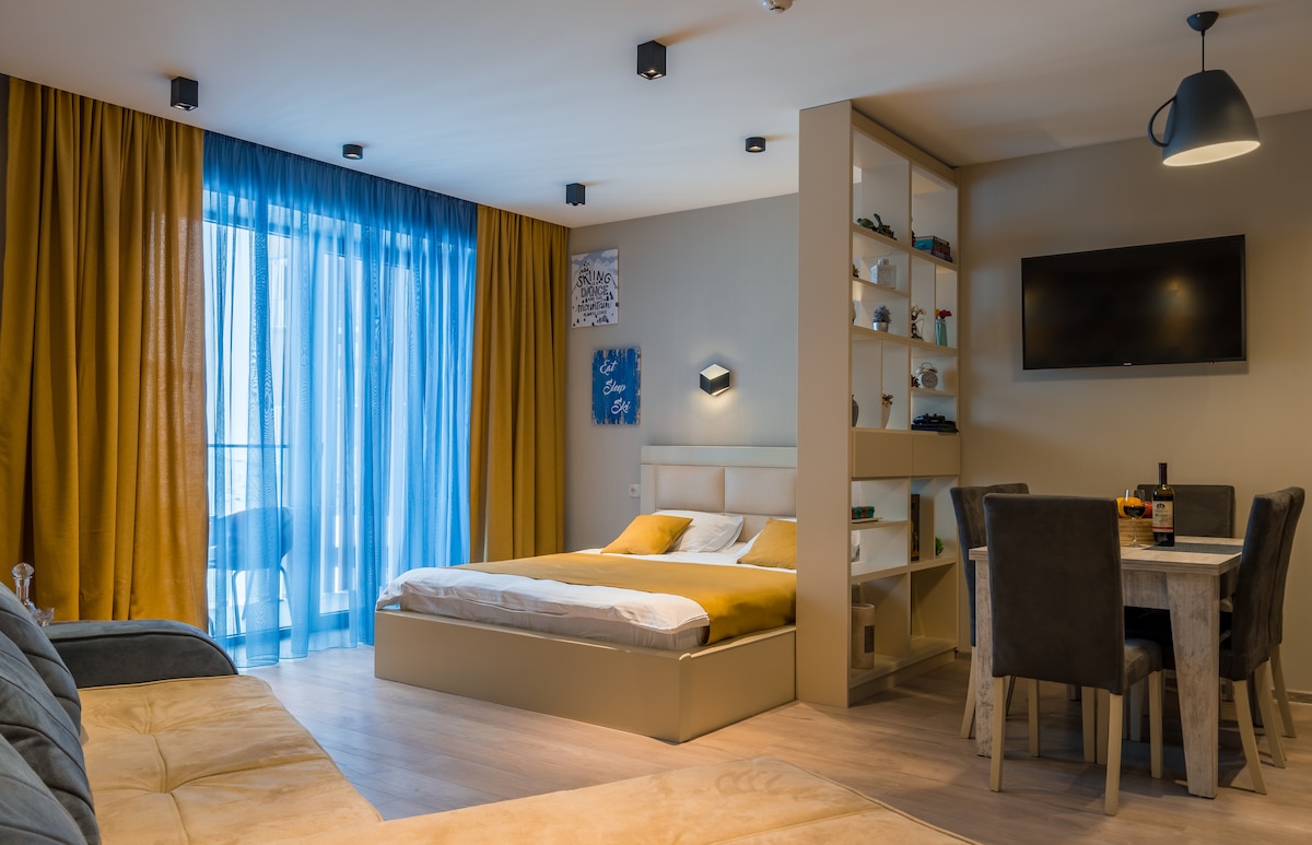 A bright living area features a spacious bed with a soft blanket, complemented by a sleek wall-mounted TV. A dining table with four chairs is positioned nearby. Floor-to-ceiling windows are adorned with blue and golden curtains, leading to a private balcony.