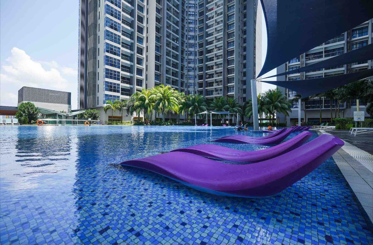 A vibrant swimming pool area features a mosaic blue tiled surface, surrounded by palm trees and modern buildings. Purple lounge chairs are arranged along the pool's edge. The area is well-lit by natural sunlight, creating a refreshing outdoor space for relaxation.