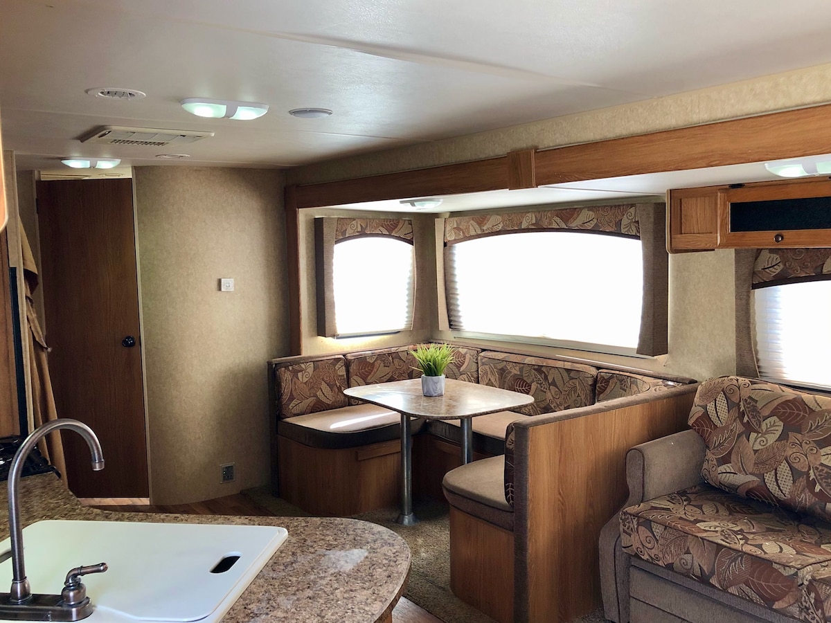 The interior of the RV features a cozy seating area with curved upholstered benches around a central table. Large windows allow natural light to fill the space, with soft curtains enhancing privacy. A compact kitchen area is visible, showcasing a sink and countertop.