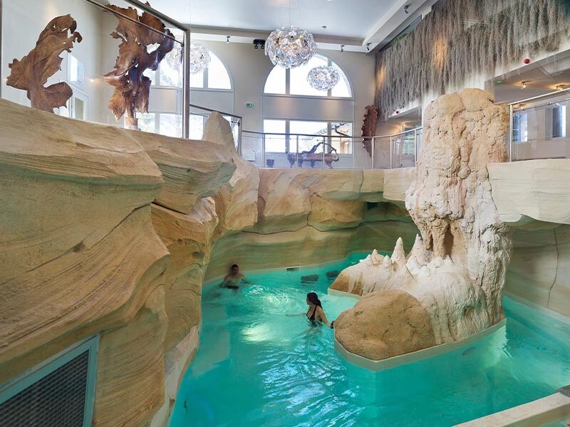 An indoor swimming pool is surrounded by natural stone-like formations, creating a serene atmosphere. Soft, turquoise water complements the elegant design, while large windows allow natural light to fill the space. Decorative light fixtures hang from the ceiling, enhancing the tranquil setting.