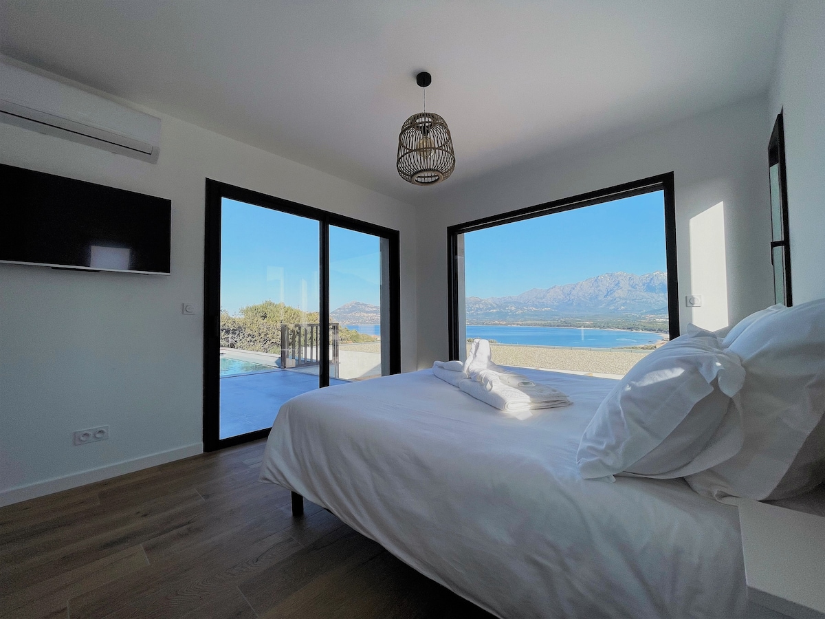 A bright bedroom features a spacious bed with freshly arranged linens. Large glass doors open to a scenic view of the landscape, while a modern wall-mounted TV adds convenience. Natural light fills the room, highlighting its minimalist decor.