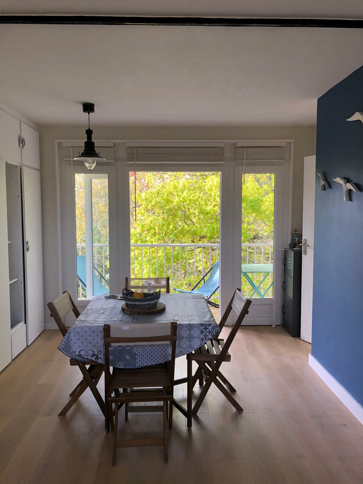 A dining area is presented with a circular table, surrounded by four wooden chairs. Large windows allow natural light to filter in while offering views of greenery outside. A decorative bowl is placed in the center of the table, adding a touch of simplicity.