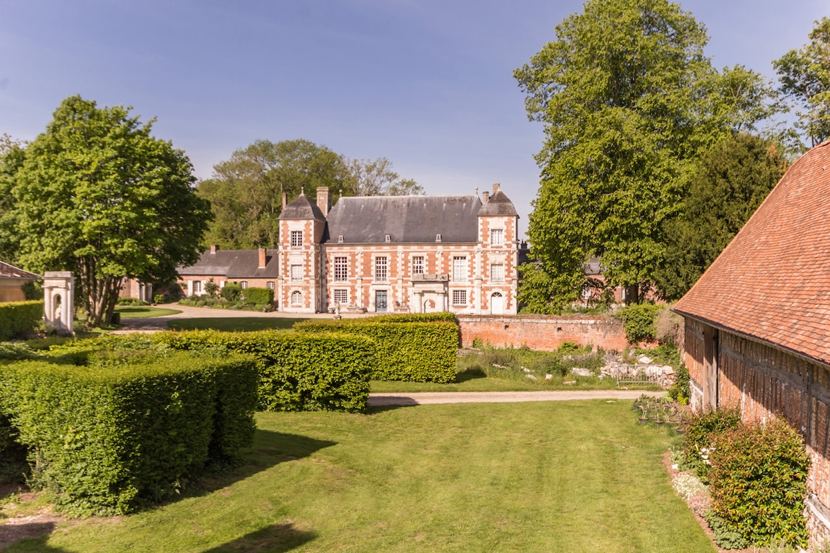 The image captures the elegant façade of a historic building surrounded by lush greenery. Well-manicured hedges frame the vibrant lawn, providing a serene setting. Trees stand tall in the background, adding depth to the picturesque landscape.