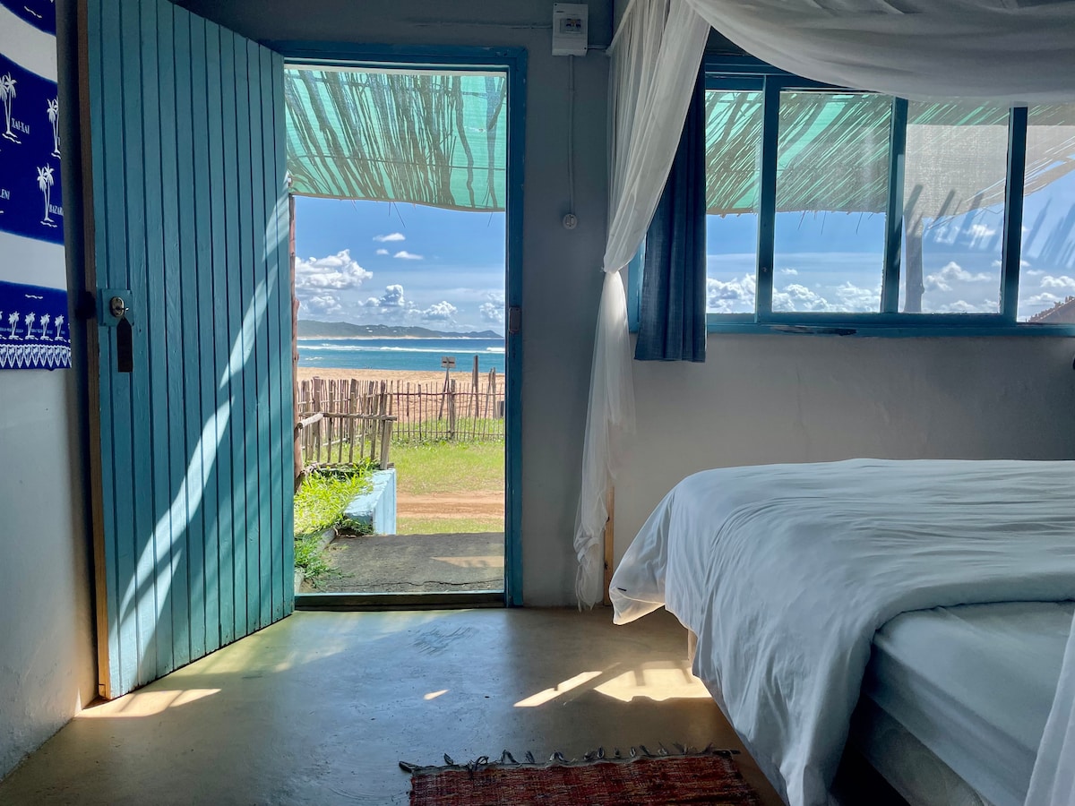 A bright room with a spacious bed is illuminated by natural light through large windows. A door framed in blue opens to reveal a view of the beach under a clear sky, enhancing the connection to the outdoor scenery.