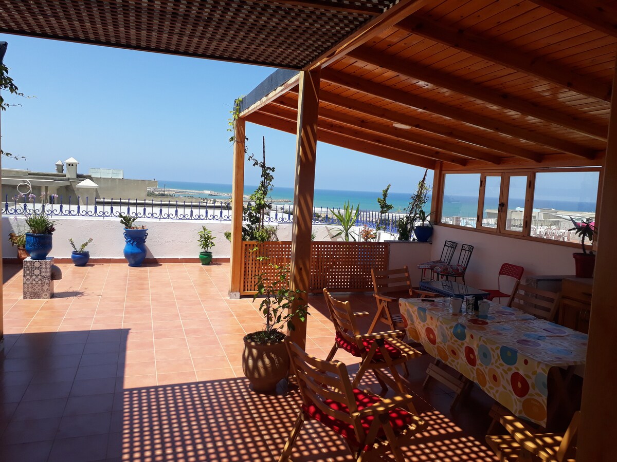 A spacious terrace features a wooden roof and tiled floor, overlooking the sea and surrounding area. Several potted plants are arranged around the space, while a large table with a colorful tablecloth is set up for dining. Wooden chairs provide additional seating.