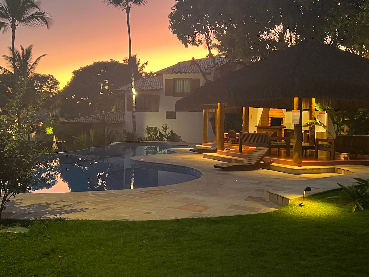 An inviting outdoor area features a large swimming pool surrounded by lush landscaping. A thatched-roof gazebo contains seating and a dining area. The scene is illuminated by soft lights, while a sunset creates a warm ambiance in the background.