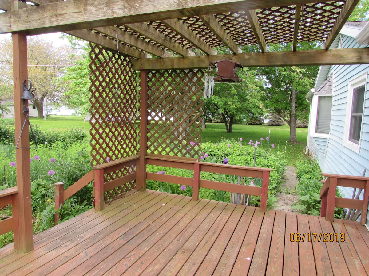 A wooden deck is covered with a lattice pergola, providing shade. Surrounded by greenery, the deck features several wooden railings and a few potted plants. In the distance, there are views of flowering plants and a lush lawn, enhancing the outdoor space.