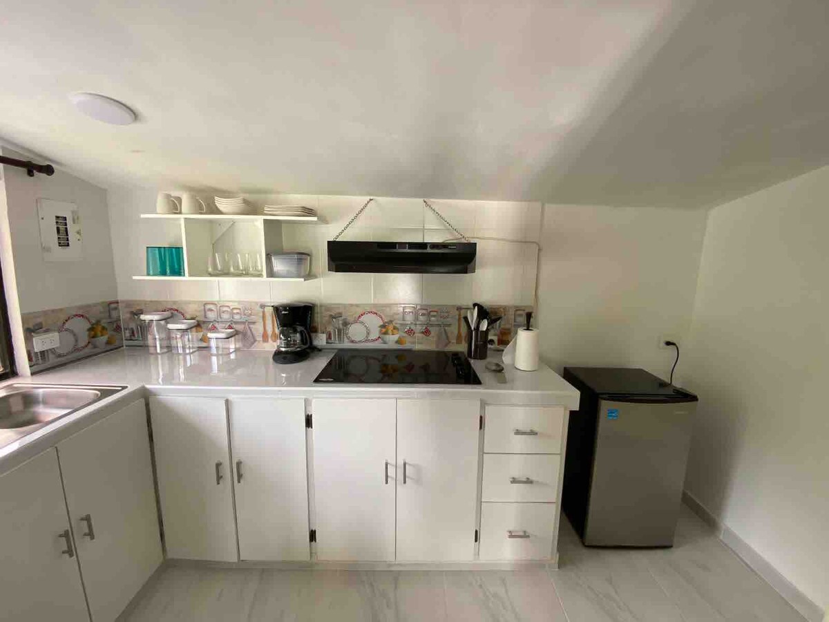 A modern kitchen space is equipped with a stovetop, coffee maker, and a compact refrigerator. White cabinetry provides ample storage, while a sleek countertop showcases various kitchen essentials. Natural light enters through a window, enhancing the functional and clean design.