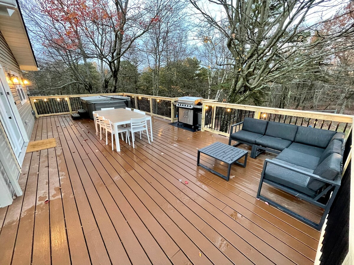 A spacious back deck features a large picnic table surrounded by white chairs, alongside a lounge seating area with dark cushions. A grill is positioned nearby, and a hot tub is visible, all set against a backdrop of trees and a soft evening sky.