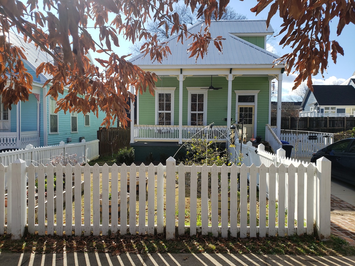 A charming historic house with light green siding is surrounded by a white picket fence. Trees with brown leaves frame the image, casting shadows on the front yard, which features a well-kept garden and a walkway leading to the entrance.