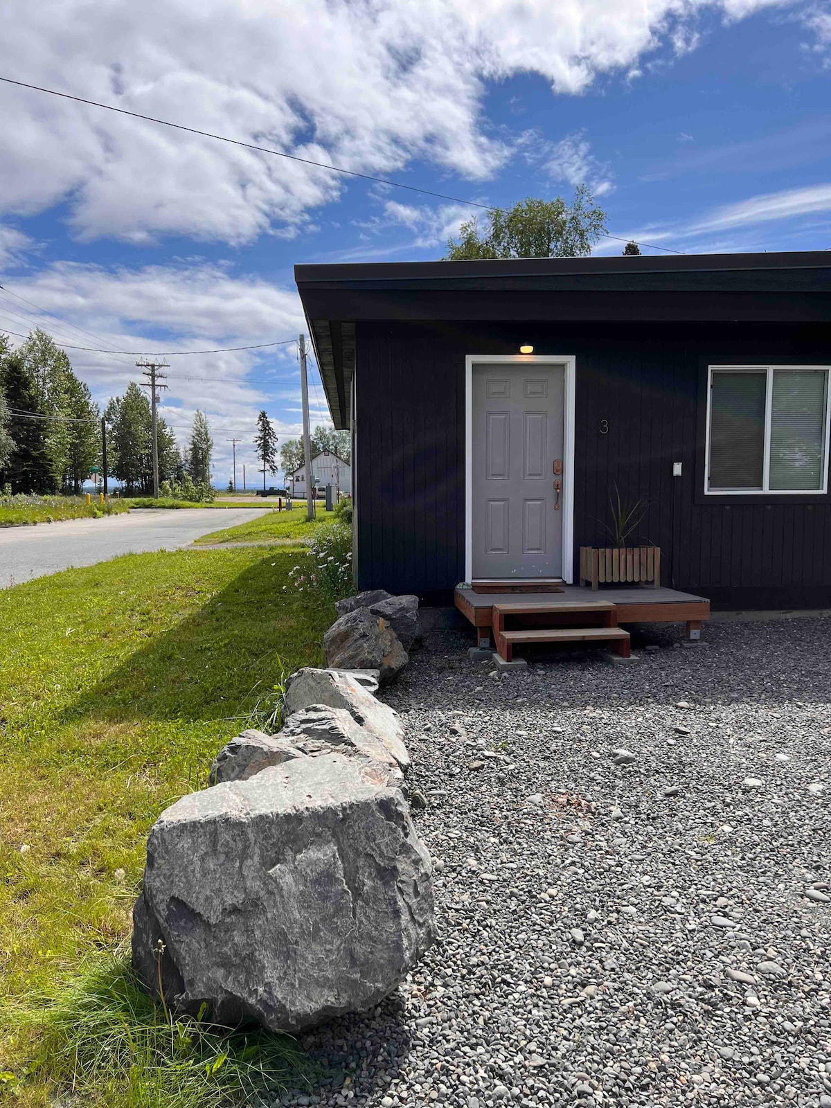 The exterior view of a modern apartment is shown, featuring a simple entrance with a wooden step leading to a white door. Large rocks line the gravel pathway. A green lawn surrounds the area, and trees and a road are visible in the background under a partly cloudy sky.