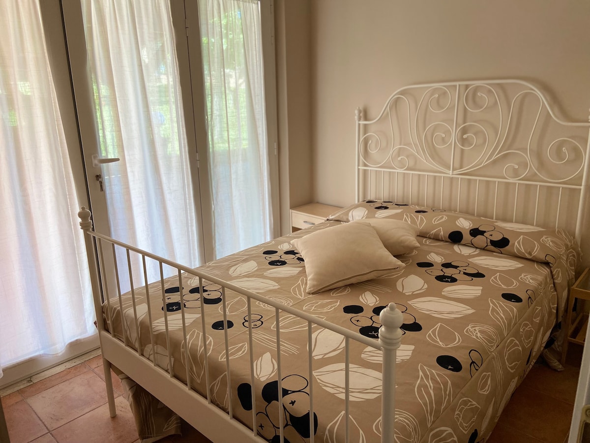 A bedroom features a white metal bed adorned with a floral-patterned bedspread. Natural light filters through sheer curtains covering glass doors that lead to a small outdoor area. A cushion is placed on the bed, complemented by a simple bedside table.