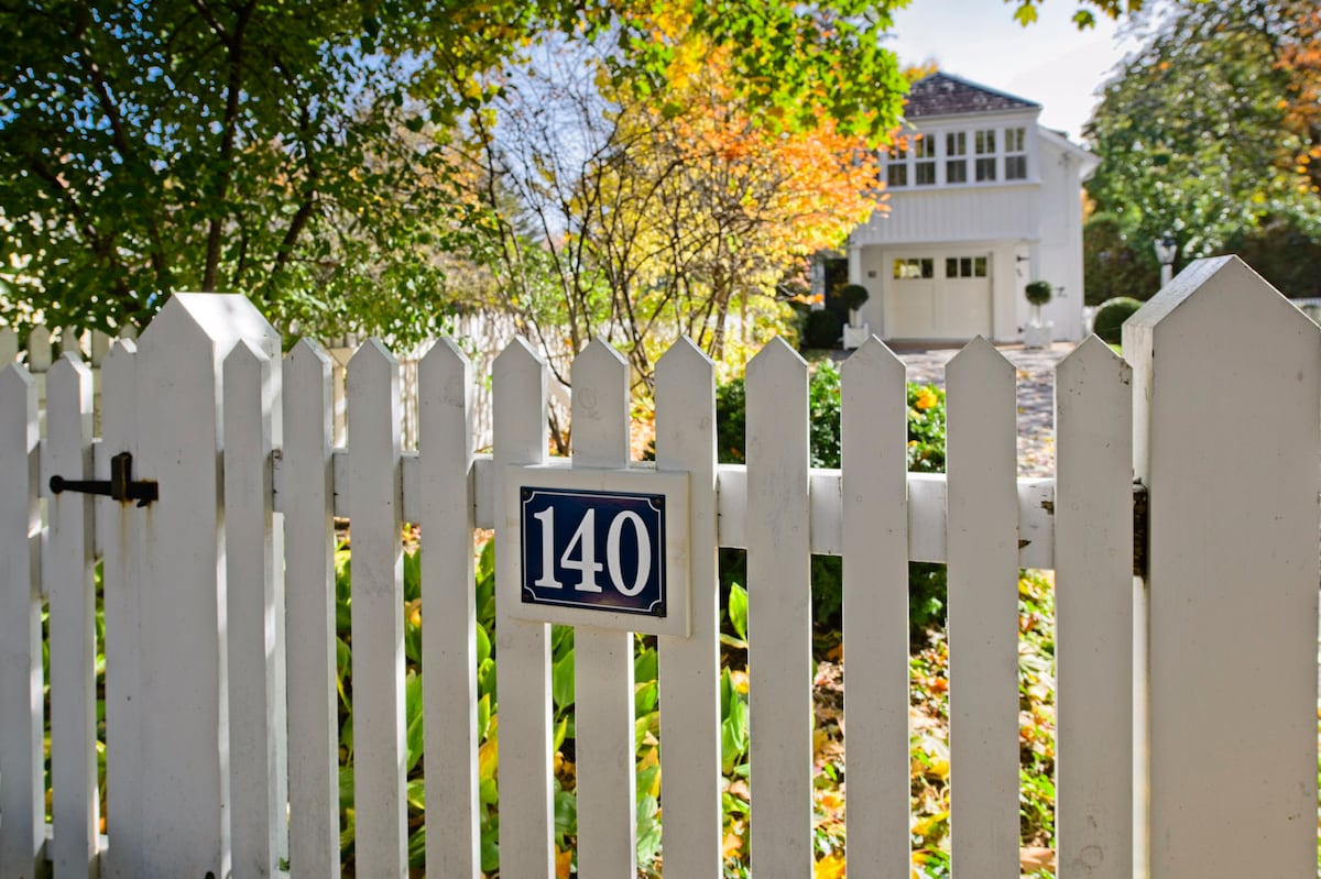 A white picket fence encloses a well-kept garden, with vibrant autumn foliage highlighting the entrance. A blue sign with the number '140' is mounted on the fence, leading to a charming house visible in the background.