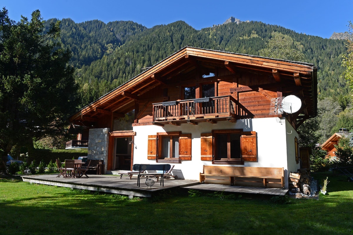 A contemporary chalet is presented within a natural setting, framed by majestic mountains. The exterior features wooden accents, large windows, and a spacious terrace with outdoor seating. A lush green lawn surrounds the building, enhancing the serene environment.