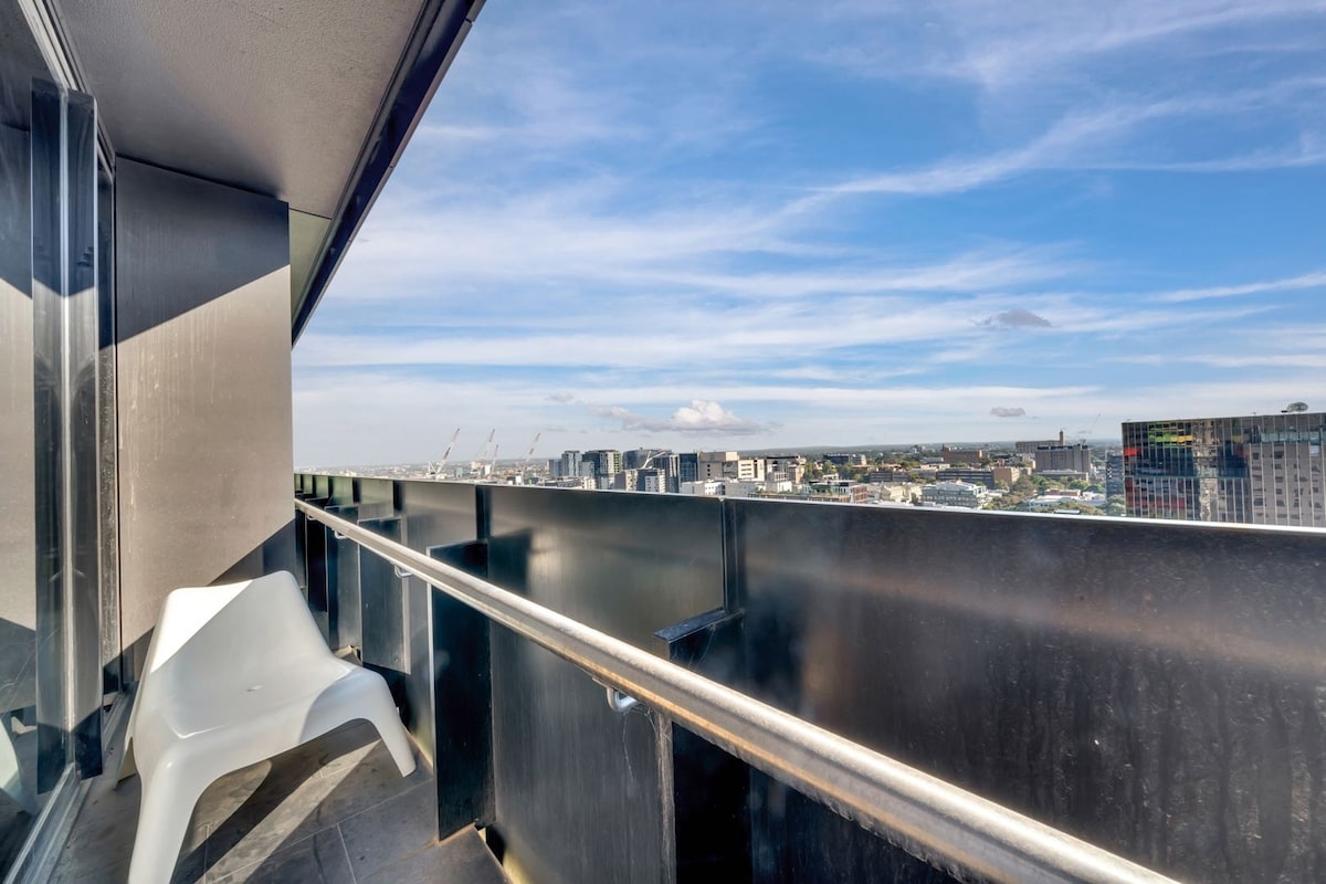 A private balcony is featured, offering expansive city views and wide-open skies. A single white chair is positioned on the balcony, complementing the modern railing and sleek design of the apartment facade.