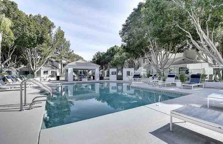 A heated glass-enclosed pool is surrounded by lounge chairs and a shaded seating area, with lush trees providing a natural backdrop. The pool area offers a tranquil space for relaxation, with a sleek design and access to nearby amenities.