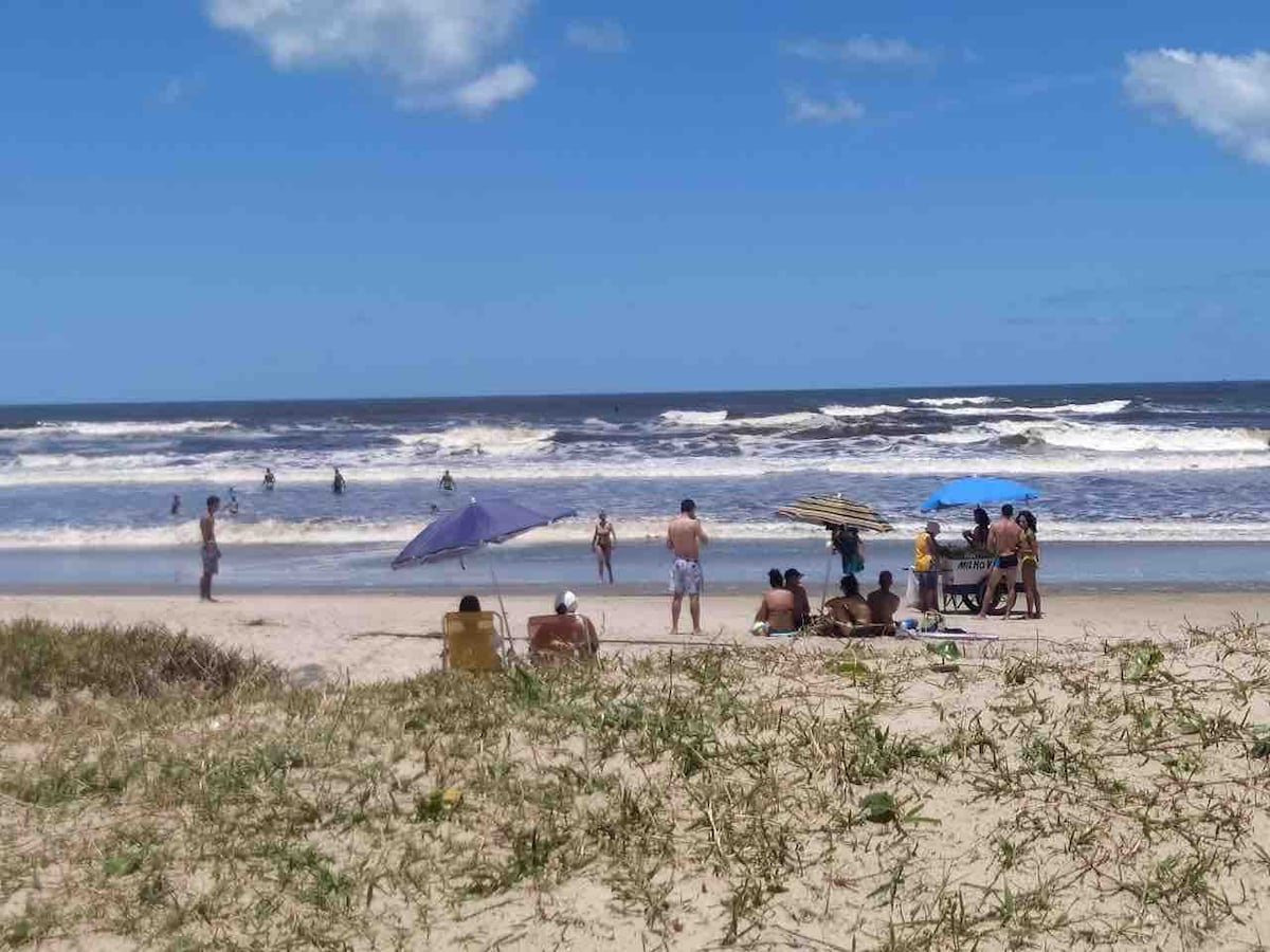 A sandy beach scene is depicted with several individuals enjoying the shore. Colorful umbrellas provide shade to some visitors, while others wade in the waves. The ocean stretches wide under a bright blue sky with a few clouds, creating a lively yet relaxing atmosphere.