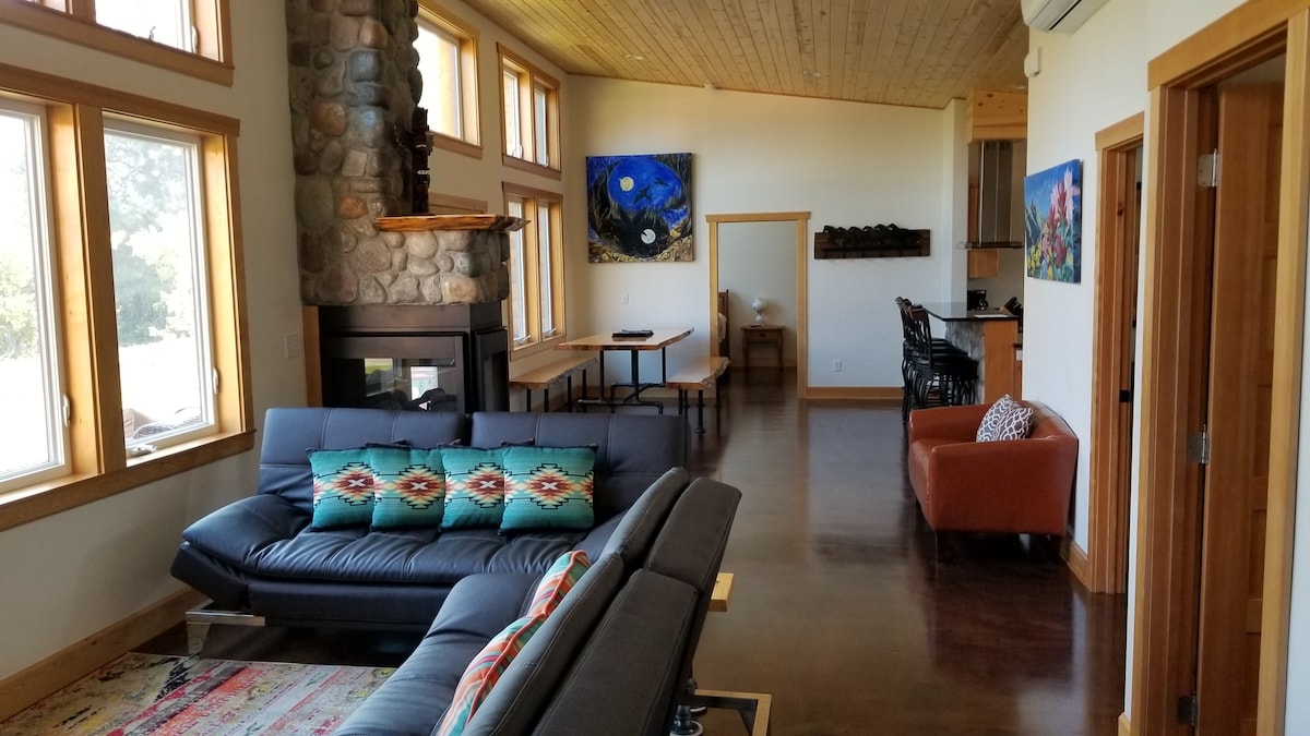 An open living area features large windows allowing natural light and views of the surroundings. A cozy sofa with decorative cushions is positioned near a stone fireplace. Original artwork adorns the walls, complementing the warm wood details throughout the space.