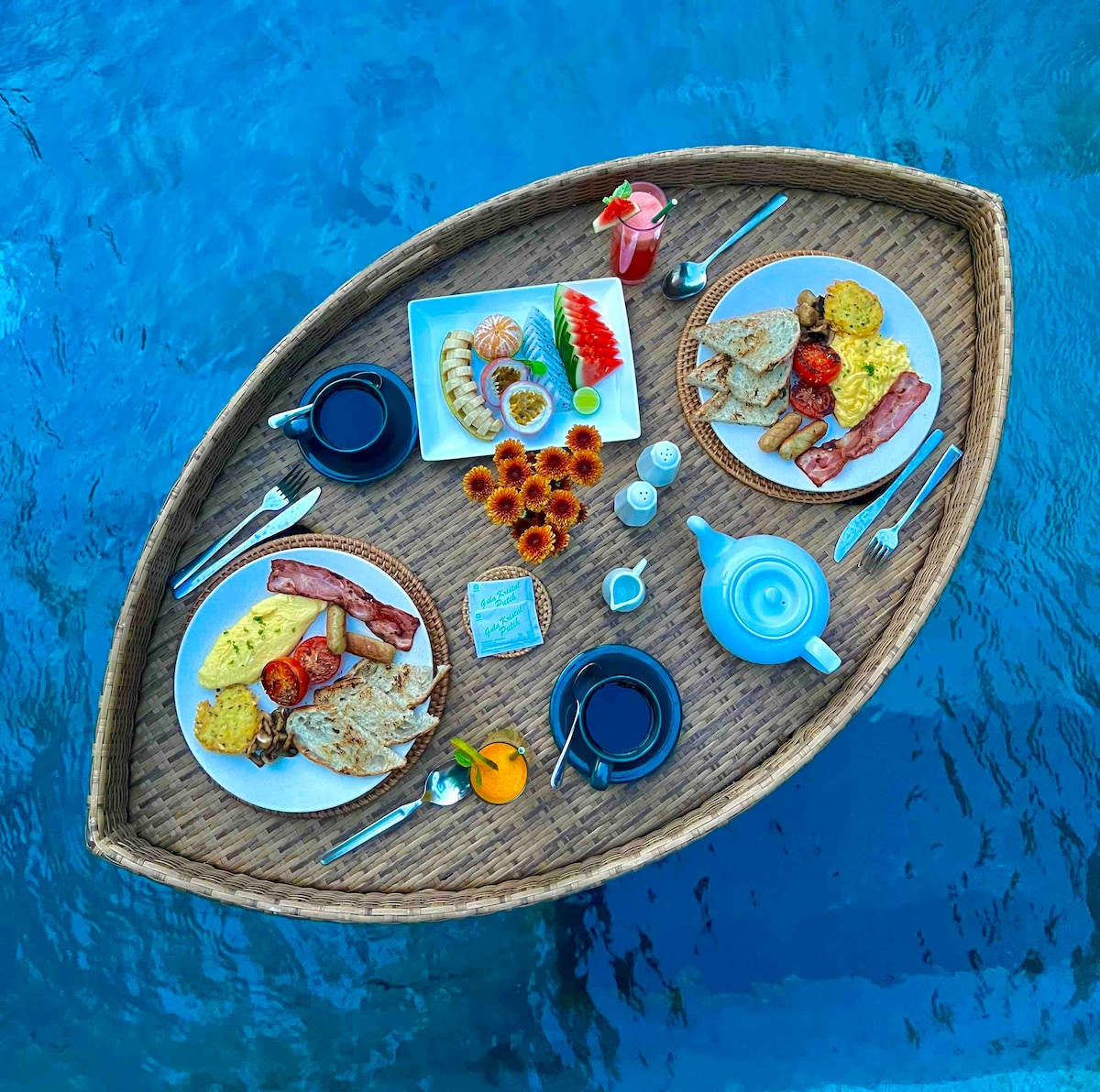 A floating breakfast tray is presented in a unique boat shape, surrounded by tranquil blue water. The tray includes two portions of breakfast with eggs, bacon, vegetables, and bread, accompanied by drinks in sleek cups and an assortment of fruits and pastries.