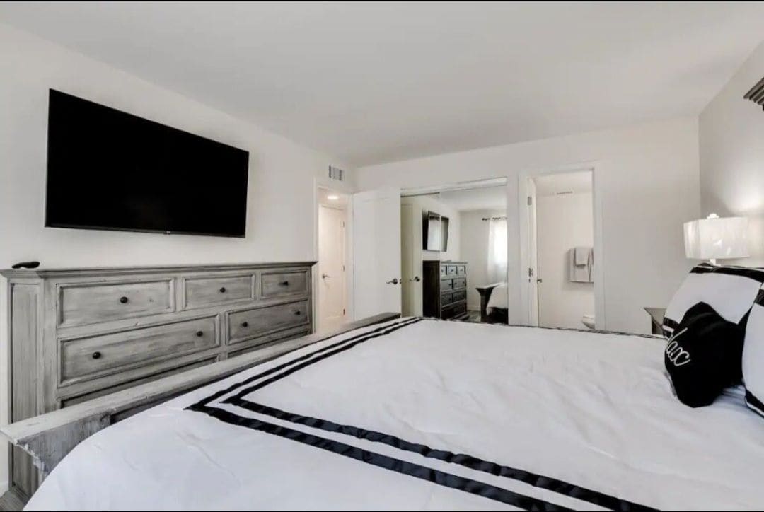 A spacious bedroom showcases a large bed with a striped blanket and decorative pillows. A flat-screen TV is mounted on the wall opposite a dresser with multiple drawers. The room features neutral tones, with ample natural light coming from a nearby doorway.