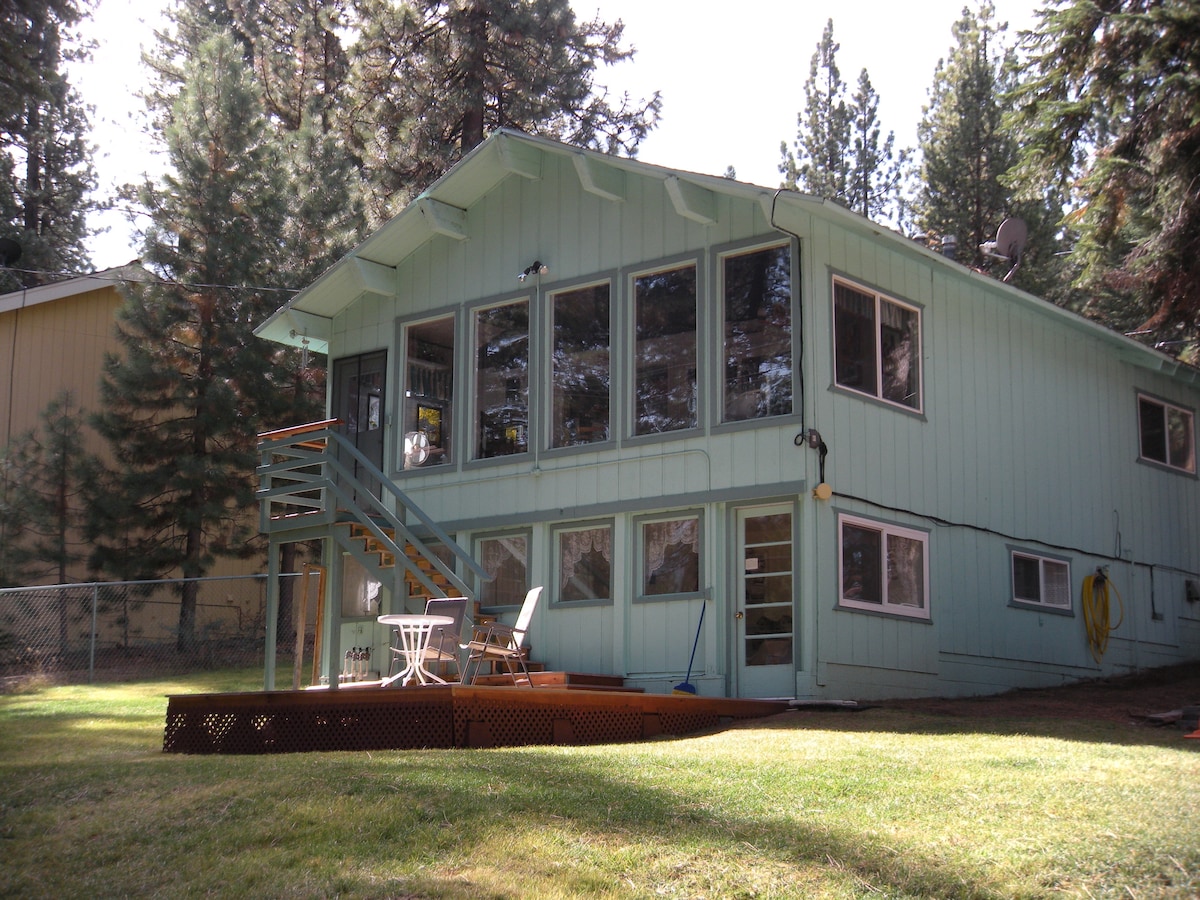 The exterior of the vacation cabin is shown, featuring a light blue façade framed by tall trees. Large windows allow for natural light, while a private deck with a small table and chairs offers outdoor seating. A fenced yard provides added privacy.