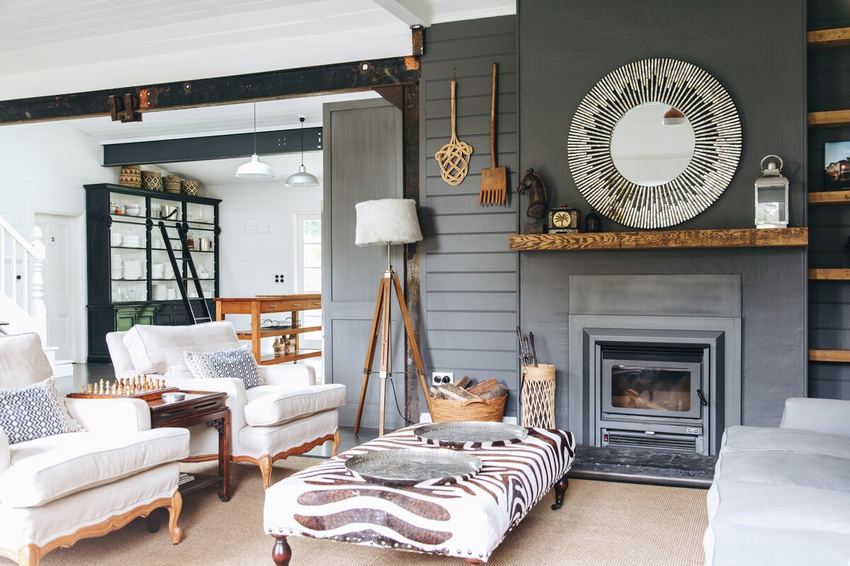 A welcoming living space features a cozy seating arrangement with two armchairs and a stylish ottoman, complemented by a contemporary mirror above the fireplace. Natural light fills the room, highlighting the elegant decor, including decorative baskets and a wooden shelf with accessories.