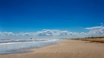 A sandy beach extends along the waterline, with gentle waves lapping at the shore under a clear blue sky. Fluffy white clouds are visible in the distance, and the coastline stretches into the horizon, providing a sense of openness and tranquility.