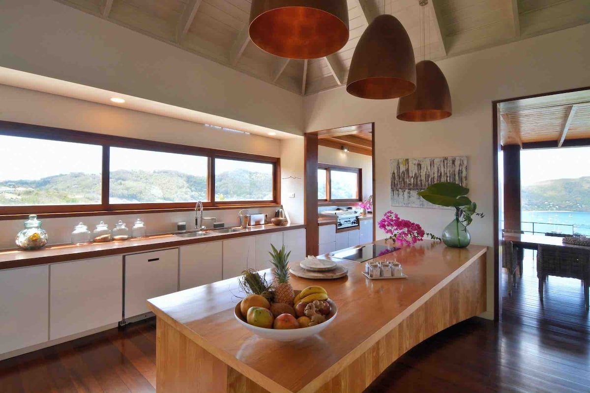A spacious kitchen is highlighted by a large wooden island at its center, adorned with a bowl of fresh fruit. Copper pendant lights hang above, providing warm illumination. Expansive windows frame views of the surrounding hills and water, creating a bright and inviting cooking space.