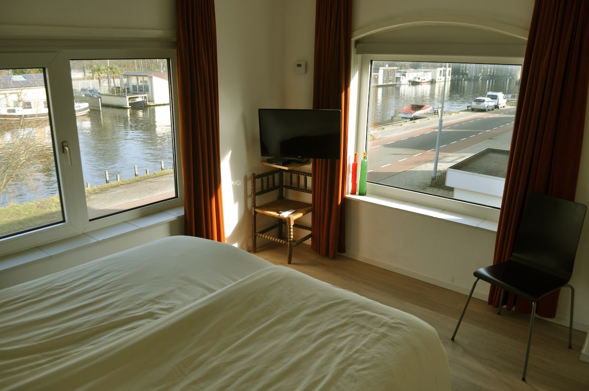A bedroom is shown with large windows offering views of a canal and houseboats. Natural light fills the room, highlighting a spacious bed with a light-colored blanket. A television is mounted on the wall, and a chair is placed near the window.