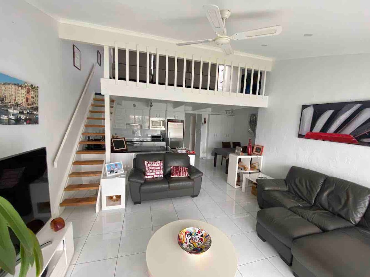 An open-plan living area features a cozy couch arrangement and a coffee table. A staircase leads to a lofted space. Modern kitchen appliances are visible, and light flows into the room through large windows. A decorative bowl adds a touch of color to the table.
