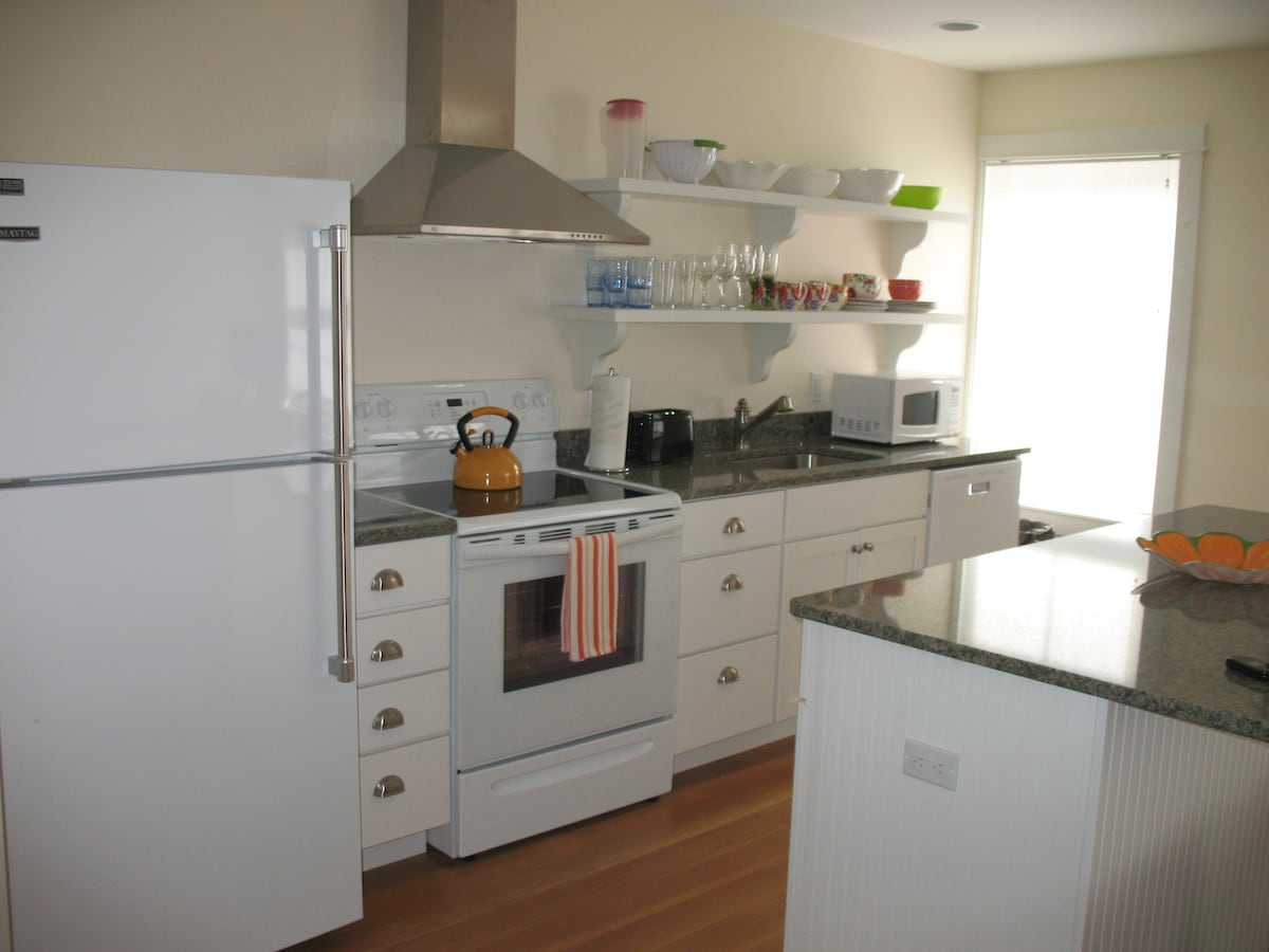 A fully equipped kitchen features sleek granite countertops and modern appliances, including a refrigerator, stove, and microwave. Open shelving displays glassware and simple kitchenware. Natural light enters through a window, illuminating the space. A bowl of fruit is placed on the island counter.