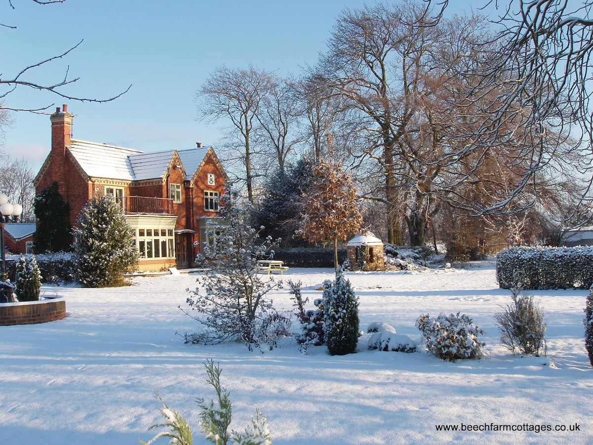 The charming cottage is surrounded by snow-covered gardens, creating a serene winter scene. Frosty trees stand majestically, and a circular stone seating area is visible in the background. The idyllic setting suggests a peaceful retreat in nature during the colder months.