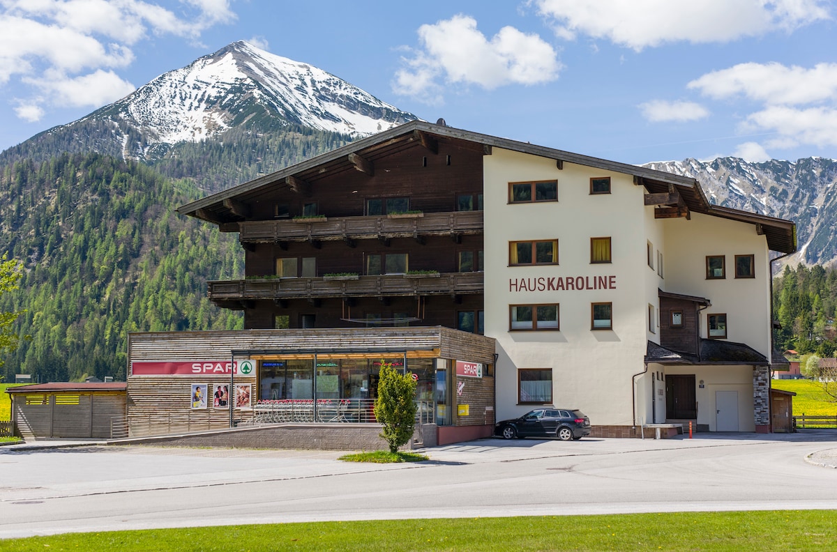 The Haus Karoline building is presented with multiple balconies overlooking the surrounding landscape. A supermarket is visible on the ground floor, with mountains rising prominently in the background. The structure features a combination of dark wood and light-colored walls, well-integrated into its natural setting.