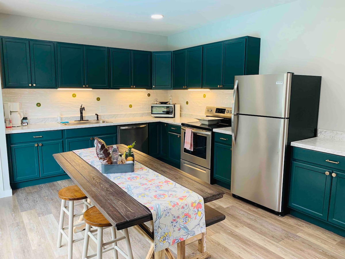 The kitchen features dark green cabinetry paired with light countertops, creating a modern aesthetic. Stainless steel appliances include a refrigerator and oven. A wooden dining table with a decorative runner is accompanied by three stools, offering a welcoming space for meals and gatherings.