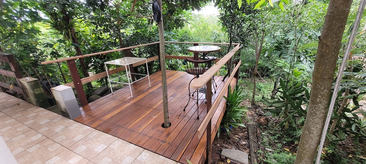 An outdoor wooden deck is surrounded by lush greenery, featuring a round table with two chairs. A shade umbrella stands at one corner, providing refuge from the sun while a natural landscape is visible in the background.