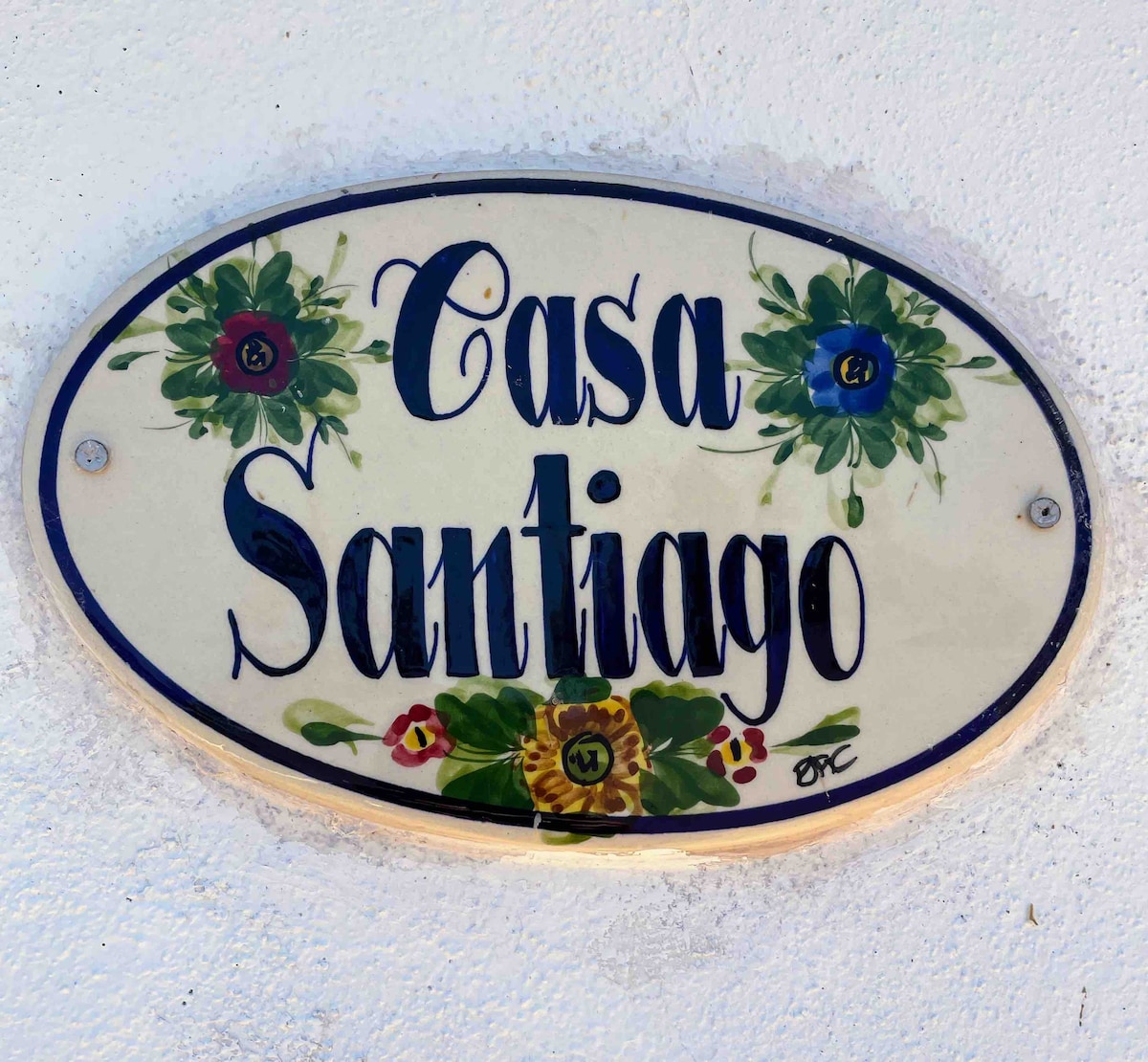 A decorative plaque displays the name 'Casa Santiago' in elegant script, surrounded by floral motifs. The sign is mounted on a light-colored wall, providing a warm welcome to guests approaching the property.