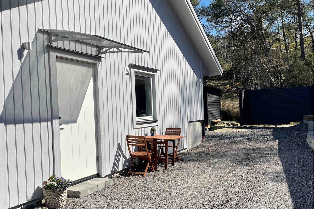 An outdoor seating area is visible, featuring a small wooden table with two chairs positioned on a gravel pathway. A wall of greenery and a solid fence can be seen in the background, providing a sense of privacy.