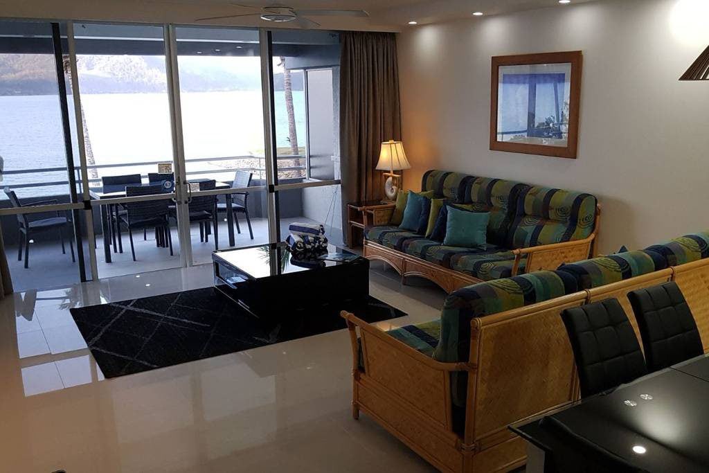 The open plan living area showcases a comfortable seating arrangement with a mix of patterned and solid cushions. Large glass doors lead to the private balcony, granting views of the ocean. A sleek coffee table sits atop a textured rug, complementing the modern decor.