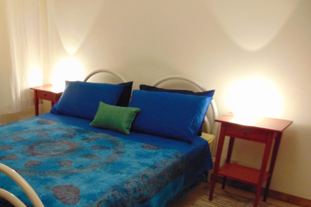 A bed is centered in the room, dressed in a blue patterned bedspread, complemented by matching blue pillows and a green accent pillow. Two red nightstands are positioned on each side, softly illuminated by bedside lamps. Light filters in through sheer curtains.